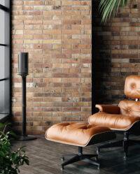 ATOM 1S Compact Oscillating Tower on its stand by a brick wall and leather chair