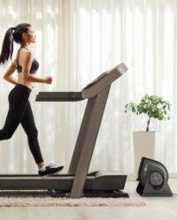 Woman in athletic clothing running on a treadmill in front of a Vornado FLY MAX and strap rote attached to the treadmill arm.
