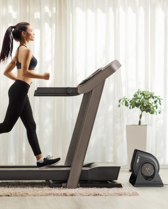 Woman in athletic clothing running on a treadmill in front of a Vornado FLY MAX and strap rote attached to the treadmill arm.